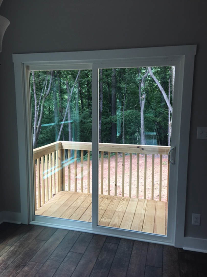 Sliding glass door opening onto a wood deck with leafy trees visible beyond, hardwood flooring inside, and wooden deck railing outside.