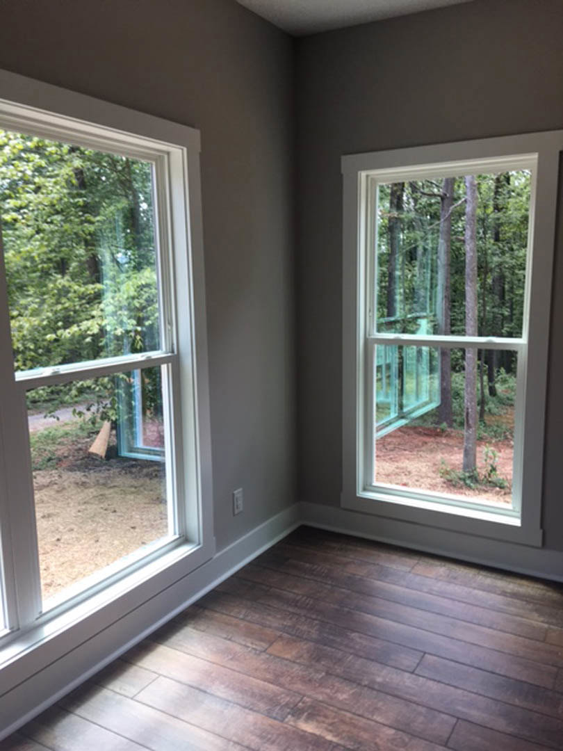 Sunlit room with two large windows, wood plank flooring, white walls, and leafy trees visible outside