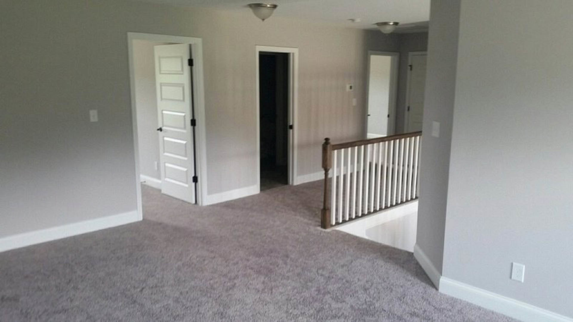 Hallway with carpeted floor, white plaster walls, wooden staircase with metal railing, multiple doors, wall-mounted light fixture, and white light switch