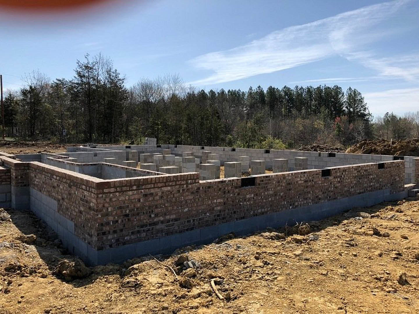 Brick foundation wall surrounded by dirt and rocks, construction site bordered by trees under a partly cloudy blue sky
