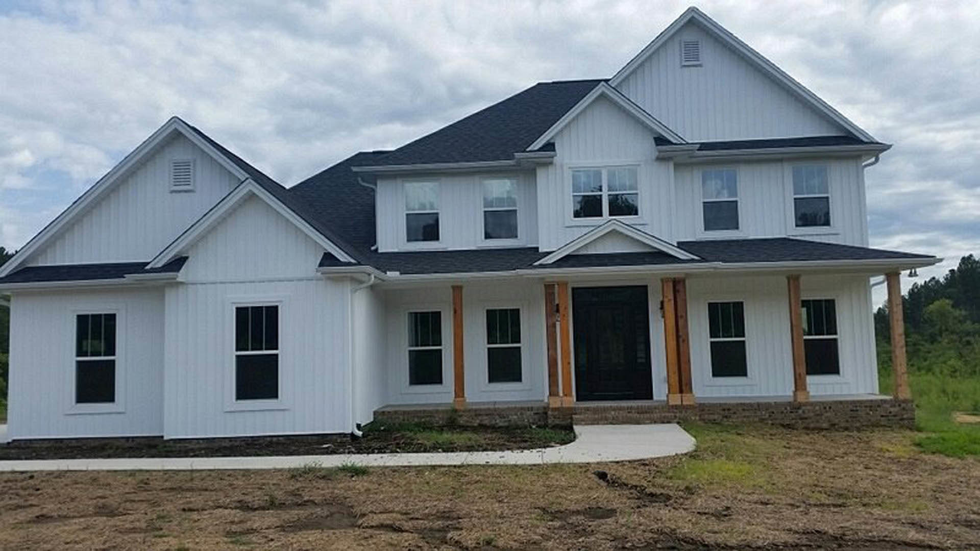 Two-story home with white siding, black front door framed in white, large windows, paved driveway, small porch, and vent on exterior wall under a cloudy sky.