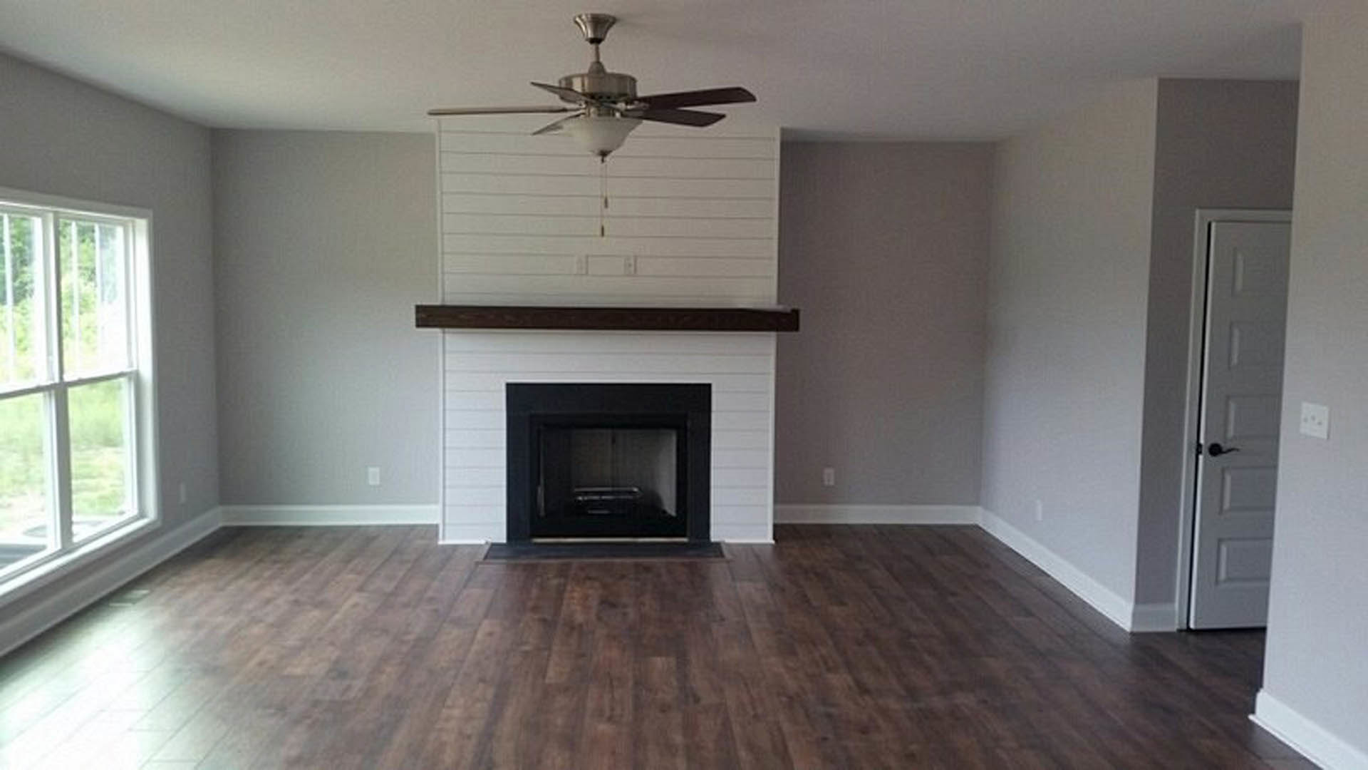 Living room with wood flooring, stone fireplace, ceiling fan with light, and large window.