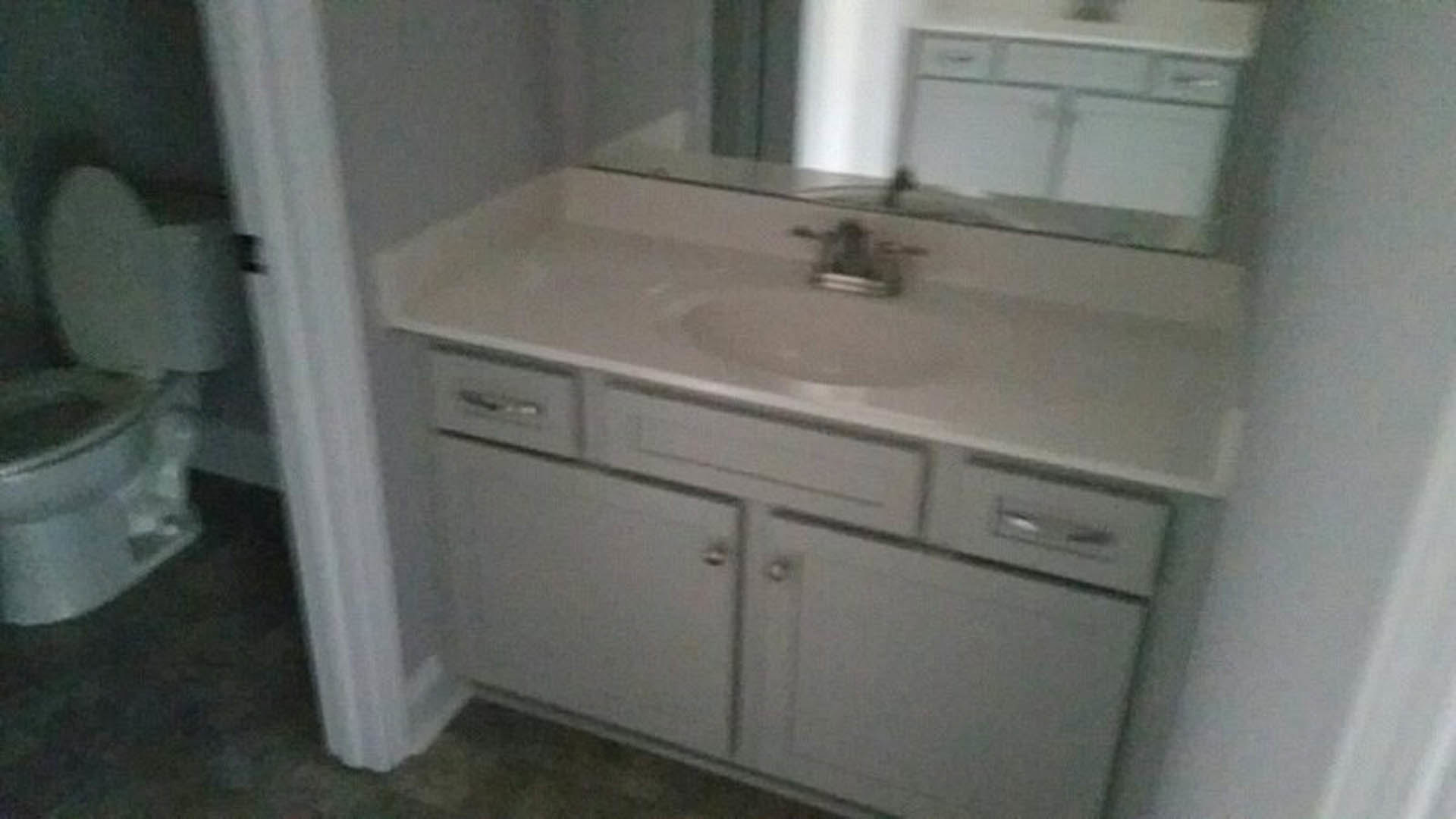 White porcelain sink with chrome faucet set in a light stone countertop, wood vanity cabinet below, tiled backsplash and wall, bathroom setting