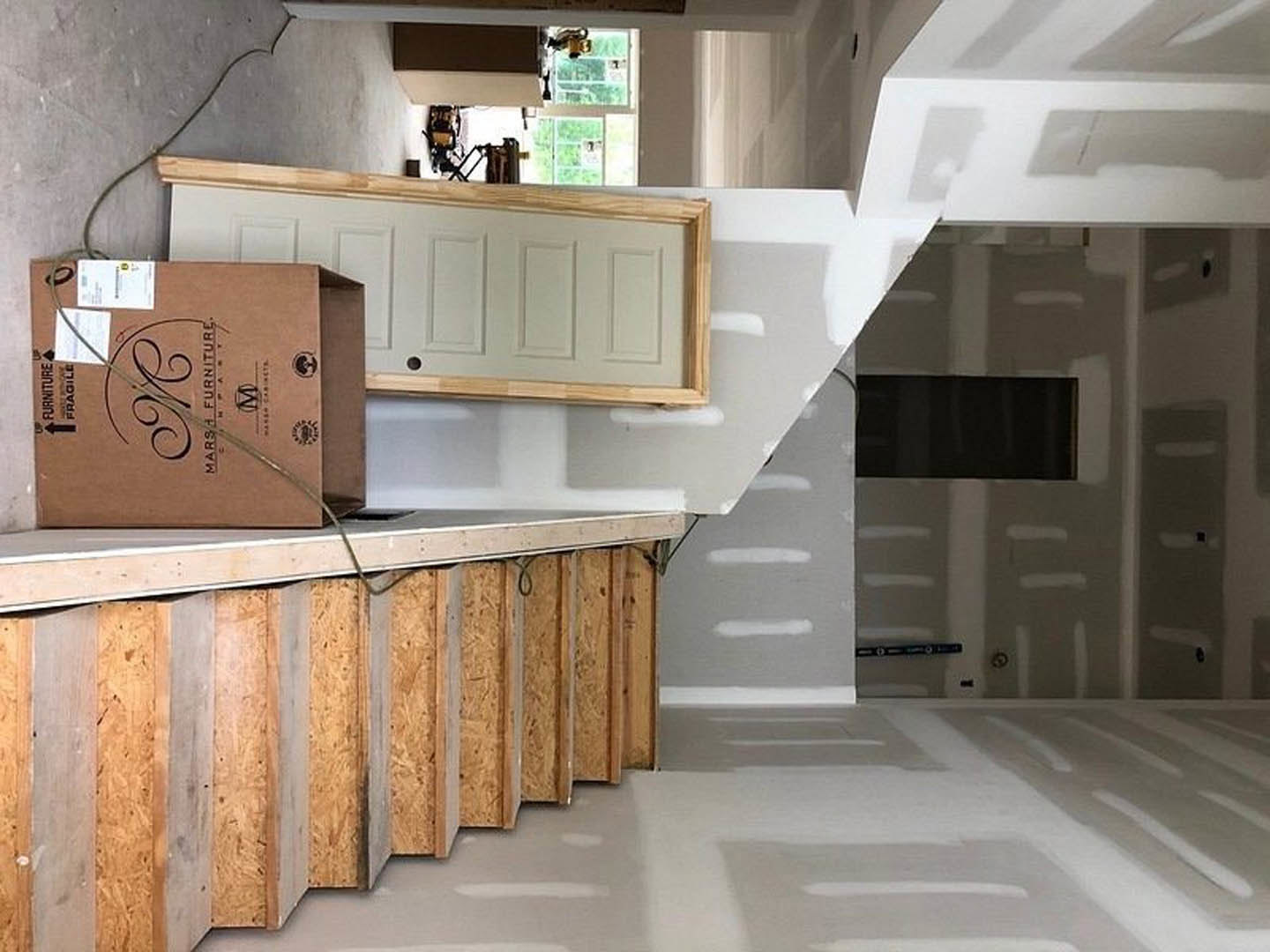 Wooden staircase with a cardboard box and cable, white door featuring a central hole, hardwood flooring, and a black rectangular object resting on a white surface.