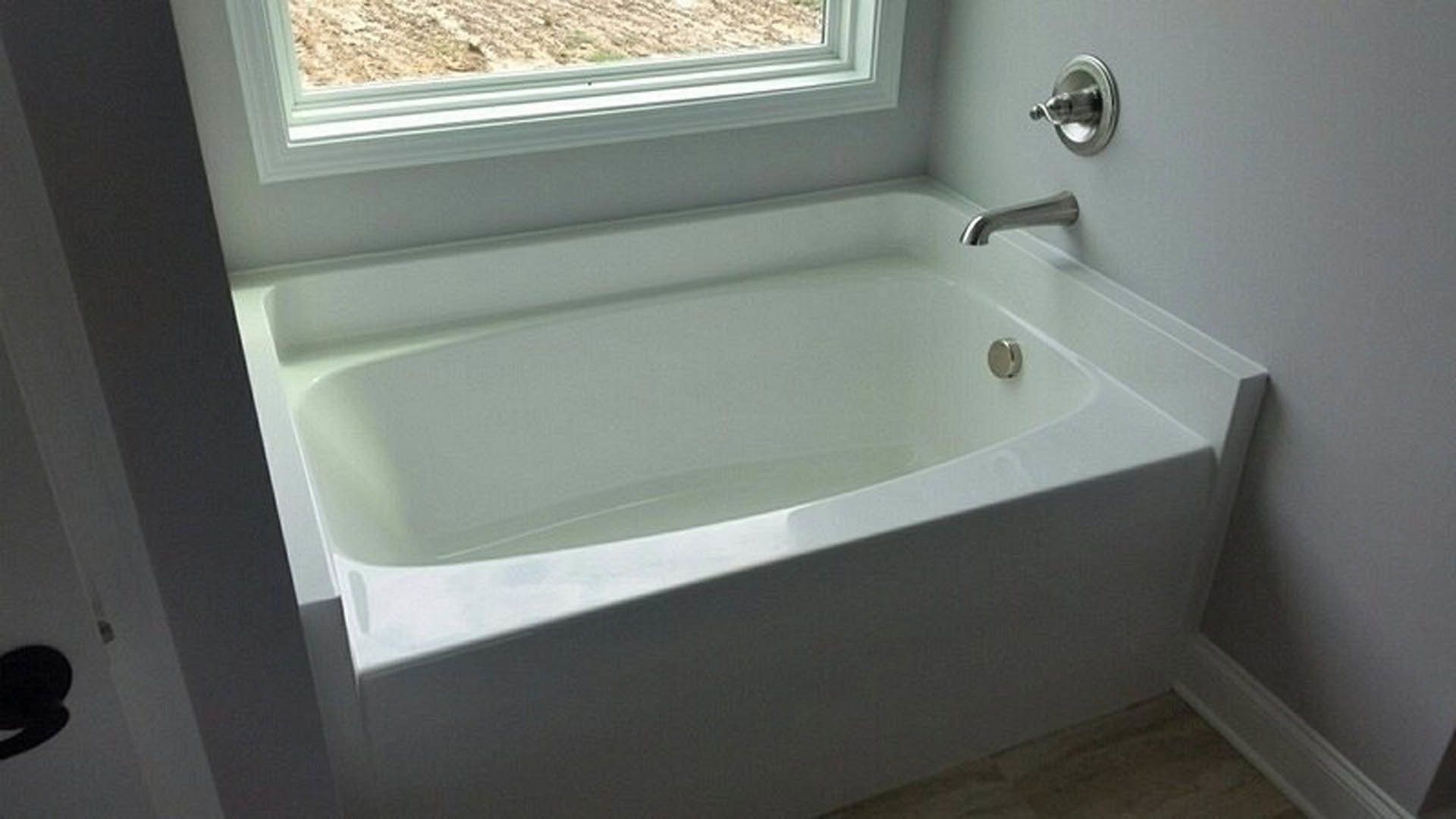 Freestanding white bathtub with chrome faucet beside large window, surrounded by light tile walls and floor, adjacent to modern sink and bathroom accessories