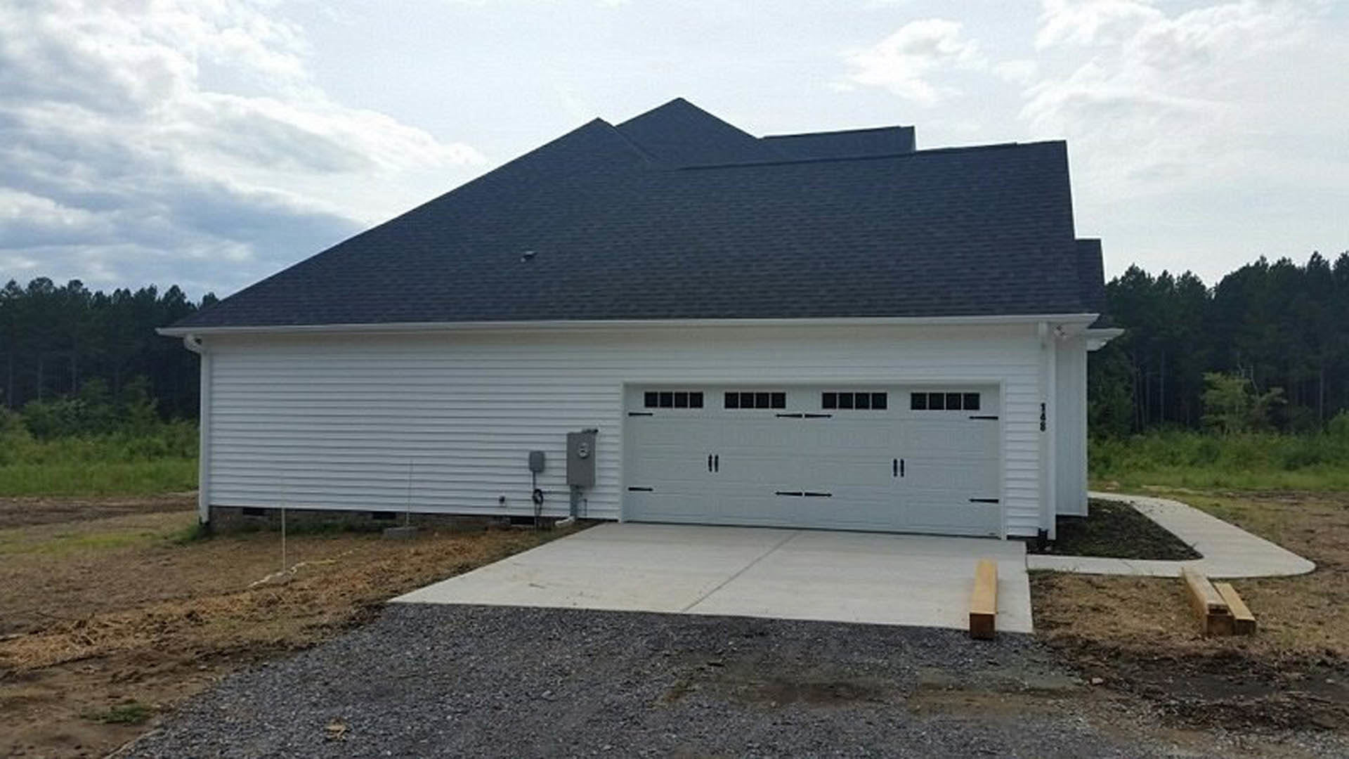 White siding house with attached garage featuring a white paneled garage door, gray shingle roof, and concrete driveway, surrounded by green lawn and mature trees under a partly