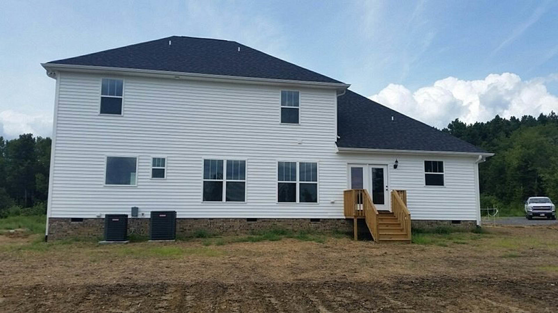 Two-story house with exterior wooden staircase and deck, large windows, white truck parked on street, dirt yard with patches of grass and rocks.