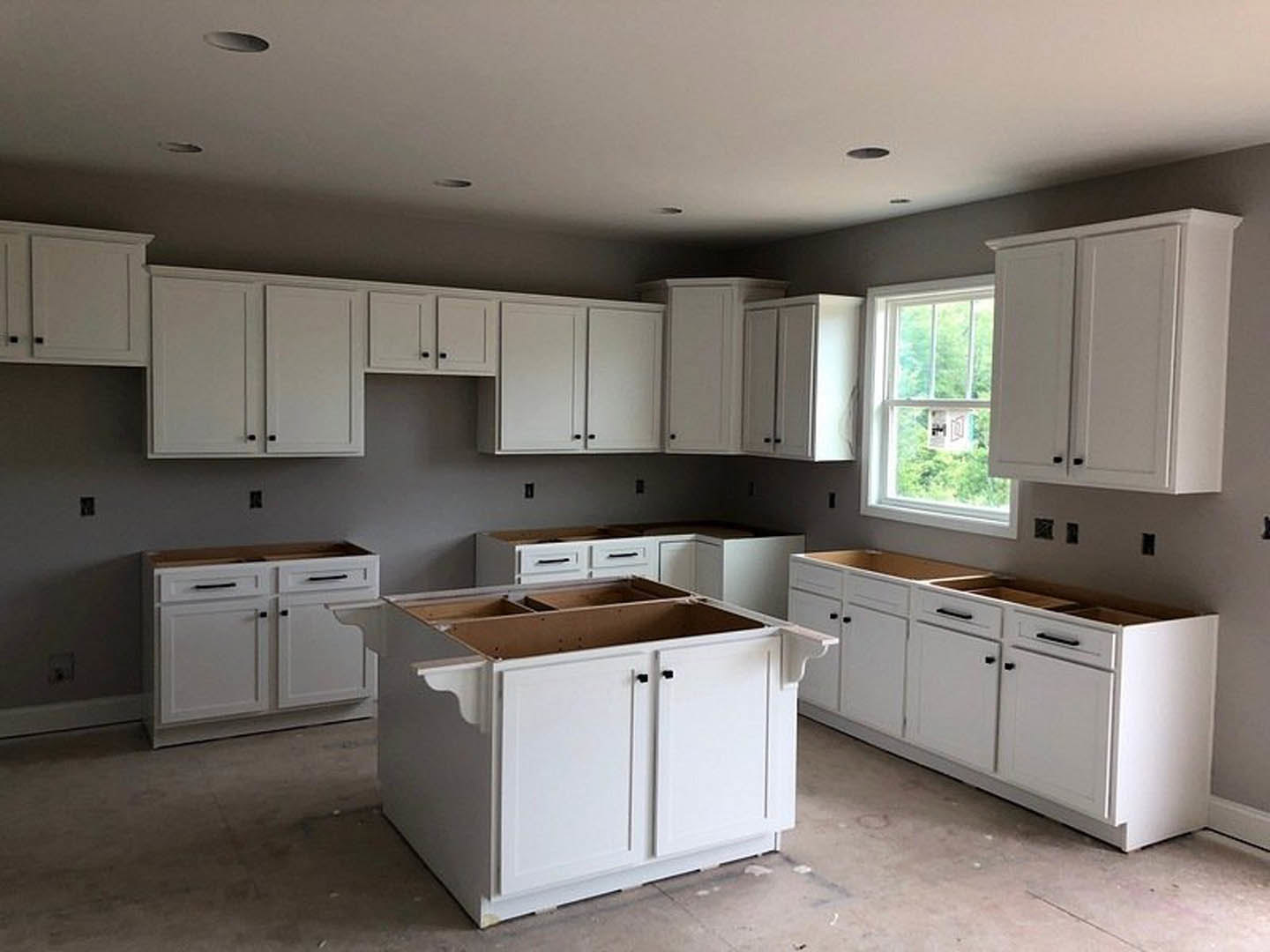 White kitchen cabinets with black handles, white countertop, stainless steel sink beneath a window with a white frame, light-colored walls, and built-in appliances.