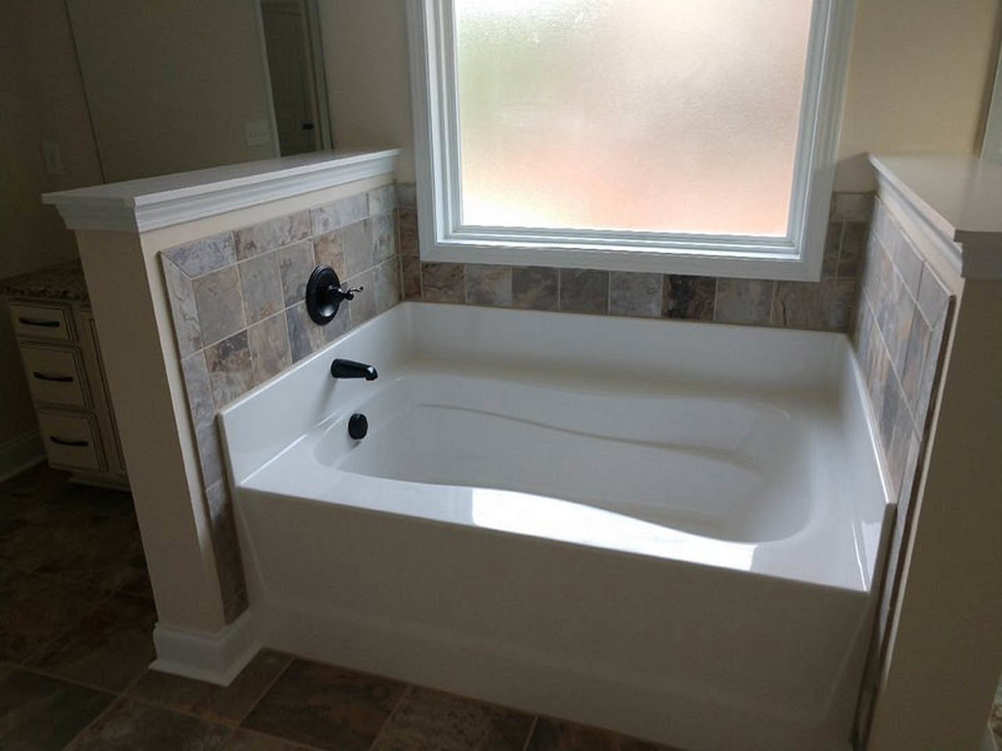 White freestanding bathtub with chrome faucet set against a light tile wall, window above, and built-in vanity drawers nearby