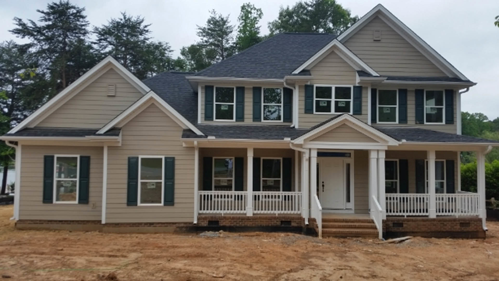 Partially built house with exposed framing, white door, brick foundation, covered porch, white-framed windows, dirt yard, and mature trees in the background