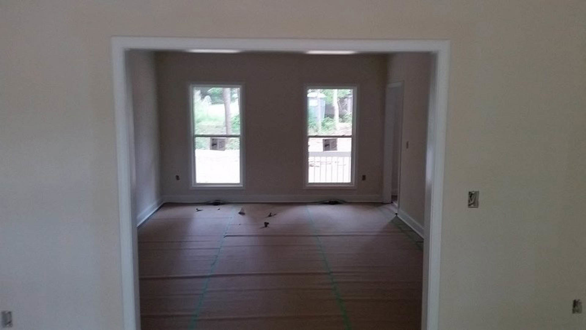 Bright room with two windows, light-colored plaster walls, wood flooring, and ceiling fixture; partial view of a white sheet with green lines and a blurry person.