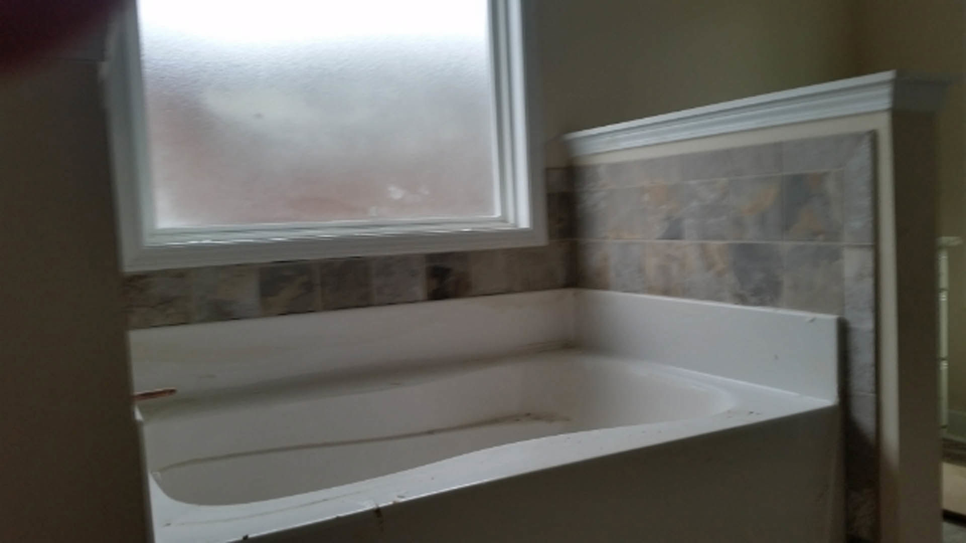 Freestanding white bathtub with chrome faucet beside large window, light gray tile walls, and built-in shower niche