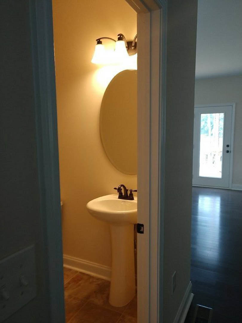 White porcelain sink with chrome faucet beneath a rectangular mirror, light gray tile walls, partial view of toilet and bathroom door