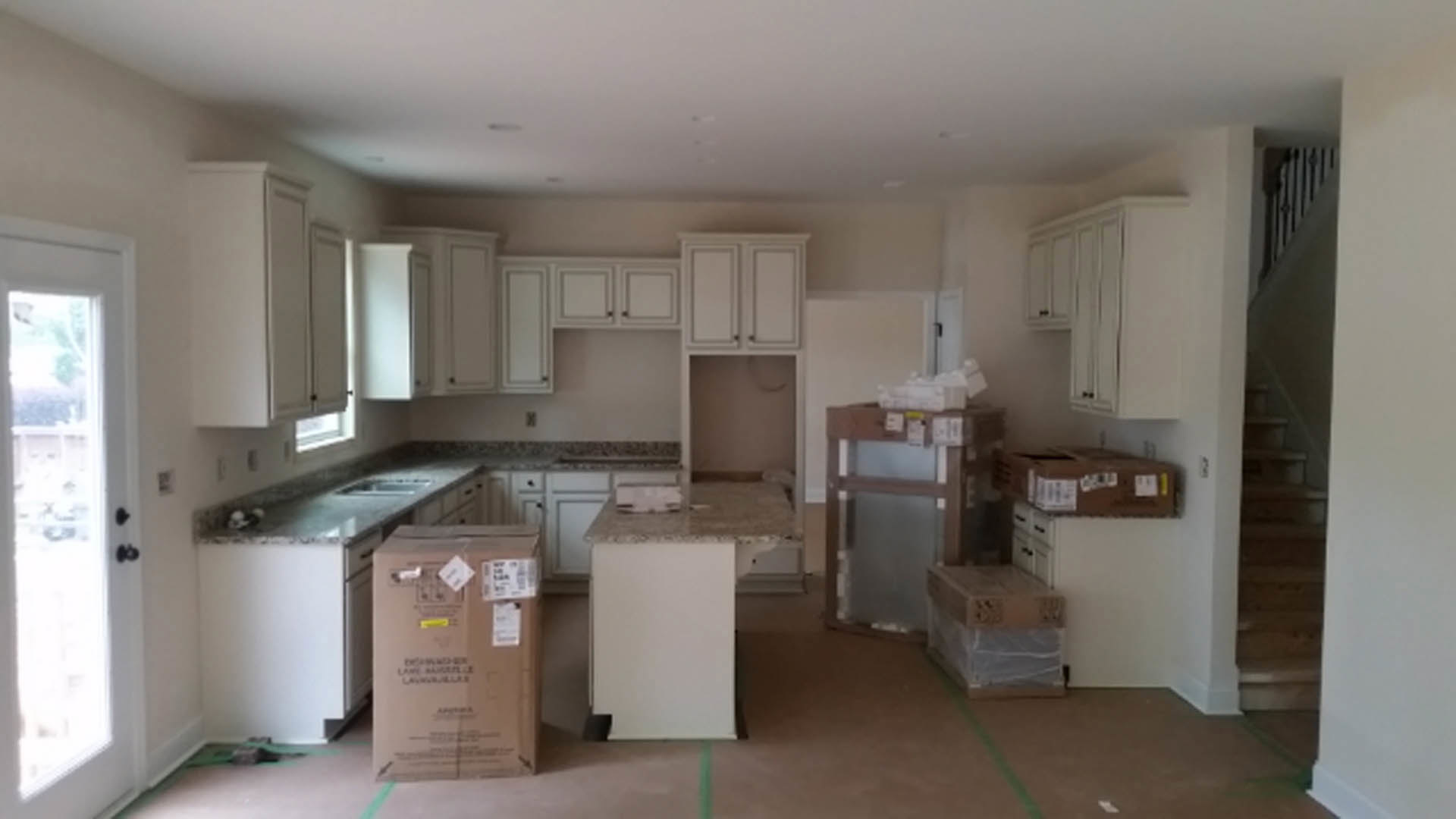 Kitchen with white marble countertop, white cabinetry, cardboard boxes with stickers and plastic wrap, wooden box with tags, white door with window, light-colored flooring, and