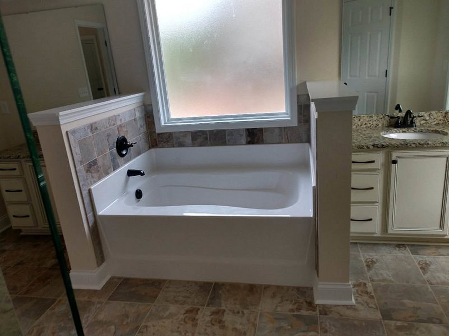 Freestanding white bathtub with matte black faucet set beside frosted glass window, surrounded by light tile walls and white cabinetry