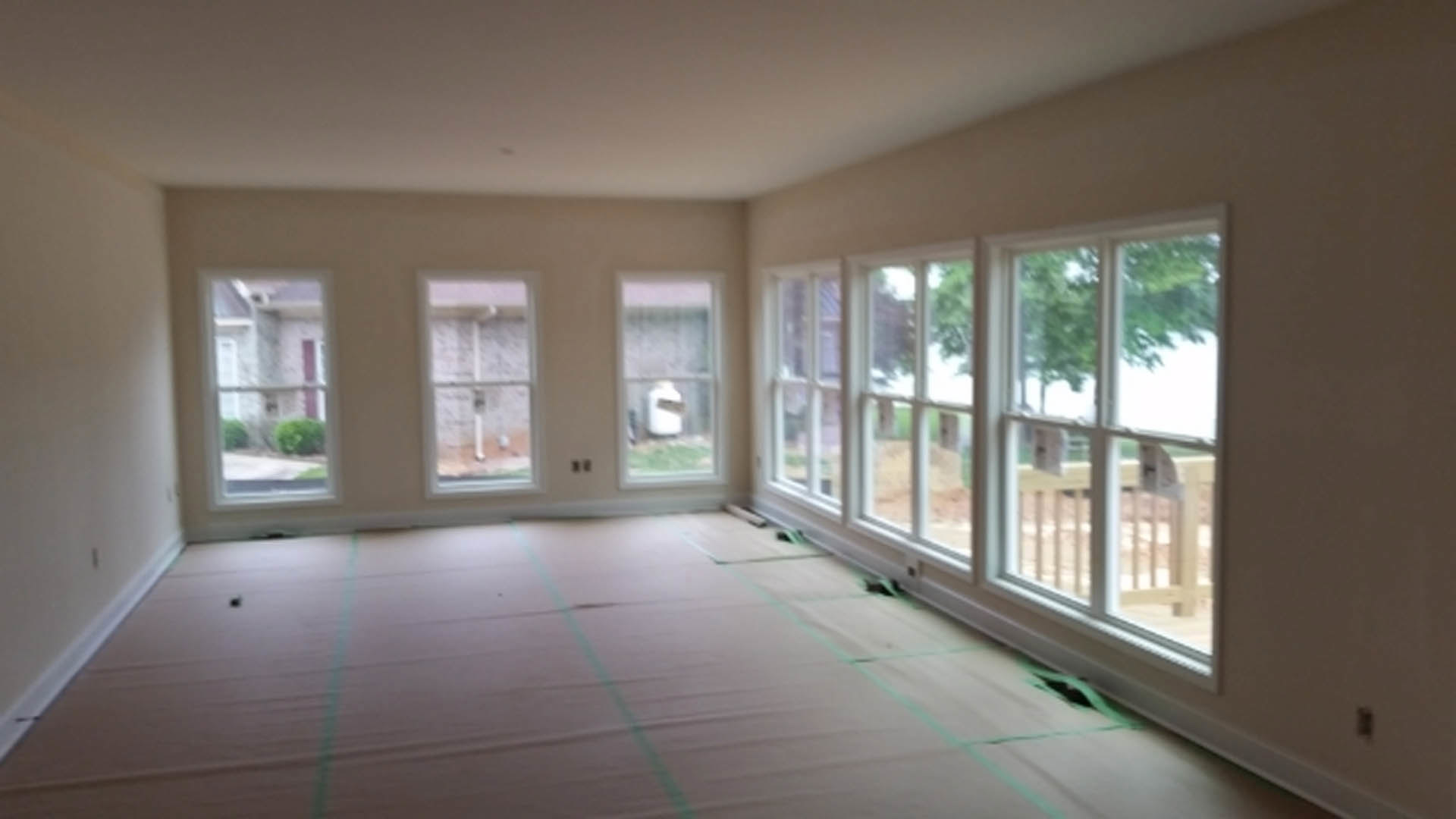 Spacious room featuring multiple large windows, light wood flooring, white walls, and views of green trees through the glass.