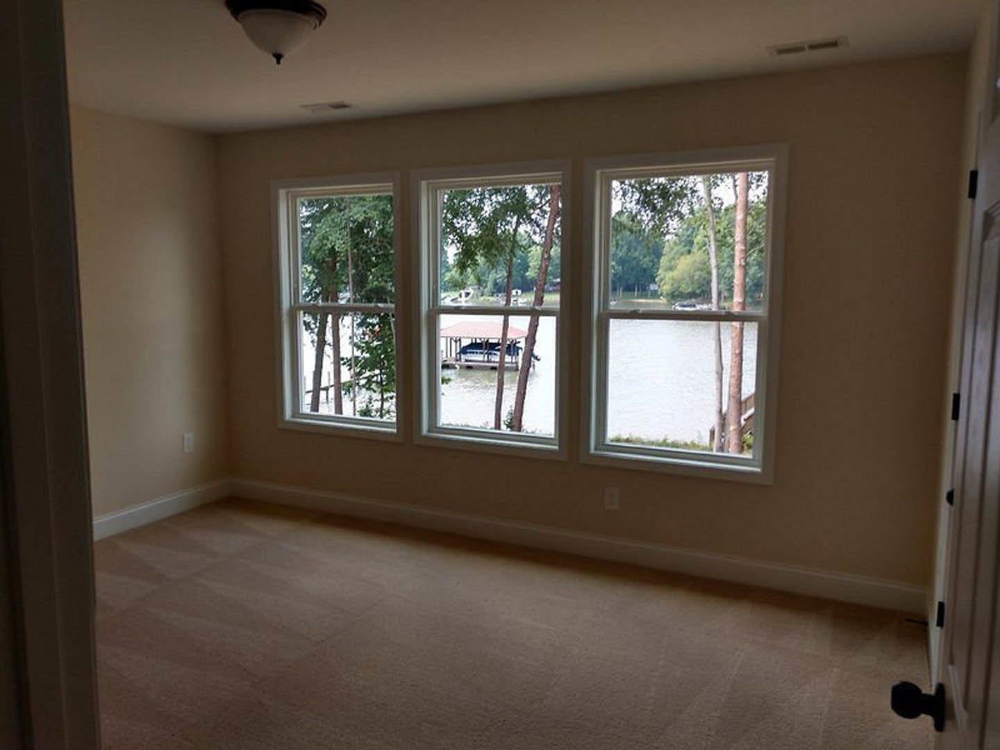 Carpeted room featuring three large windows with views of trees and water, daylight illuminating neutral walls and a modern light fixture.