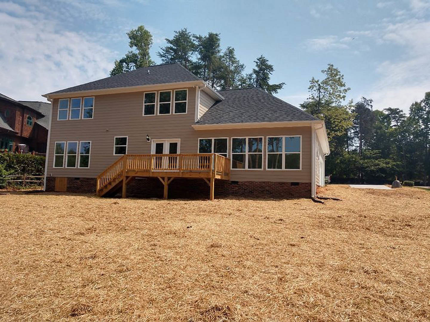 Two-story home with blue siding, white trim, wooden deck with railing, grassy yard, large windows, and gabled roof under partly cloudy sky