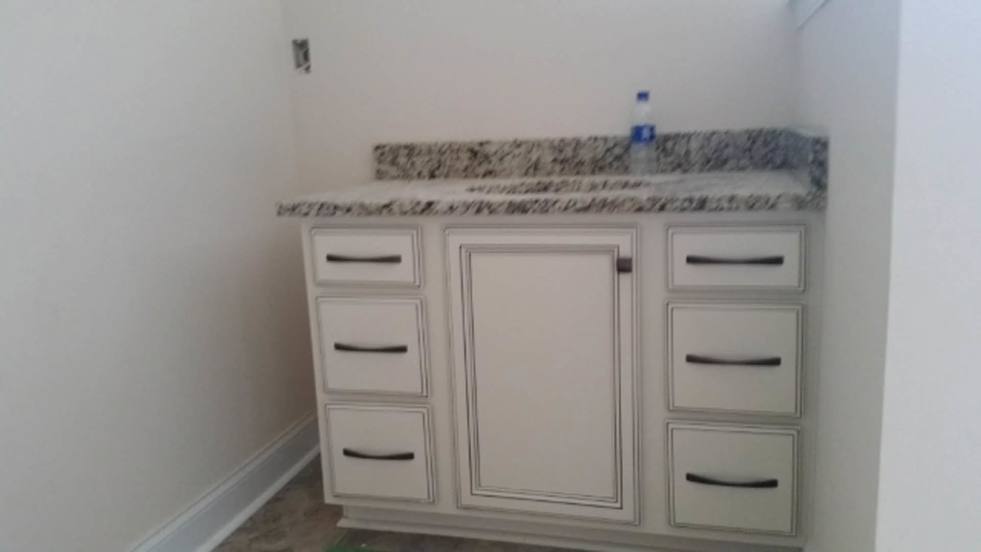 Light-colored kitchen countertop with sleek drawers, a bottle of water placed on top, white wall with black accent lines, cabinetry and blurred window in background