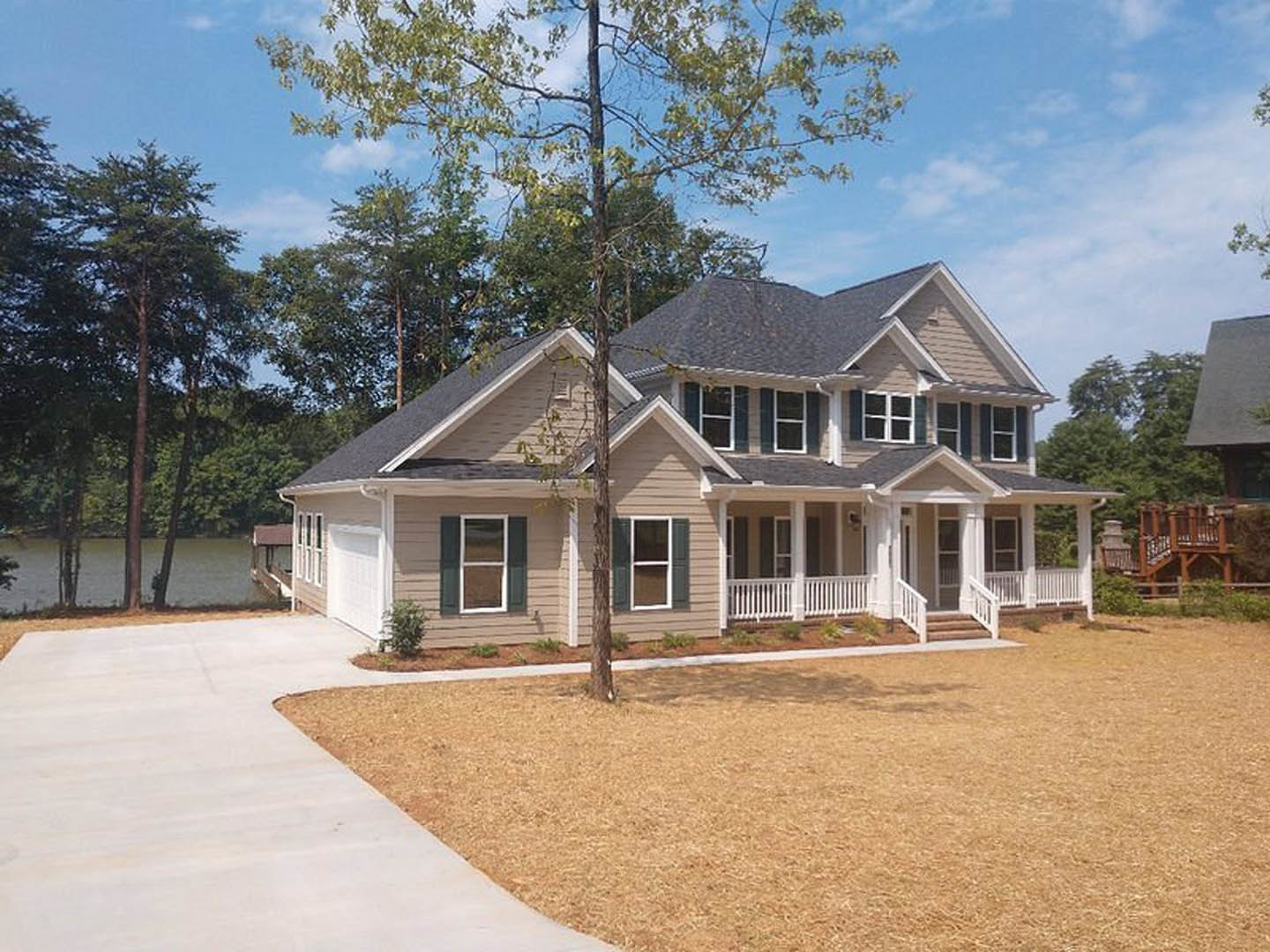 Two-story house with white-framed windows, wooden porch with railings, concrete driveway, and grass lawn bordered by mature trees