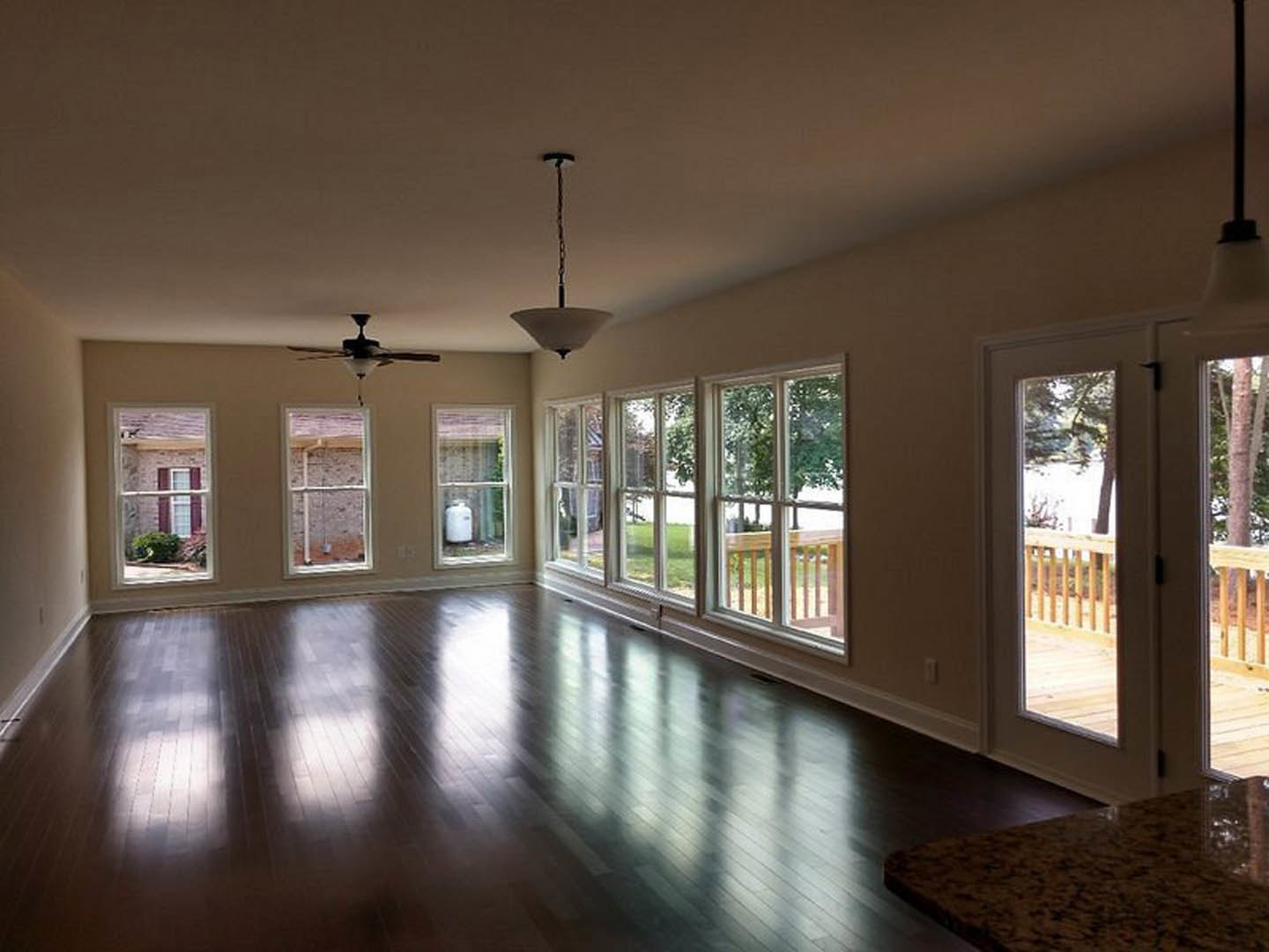 Spacious living area with laminate wood flooring, ceiling fan with light fixture, large windows providing daylight, and a view of a brick building and potted plant outside