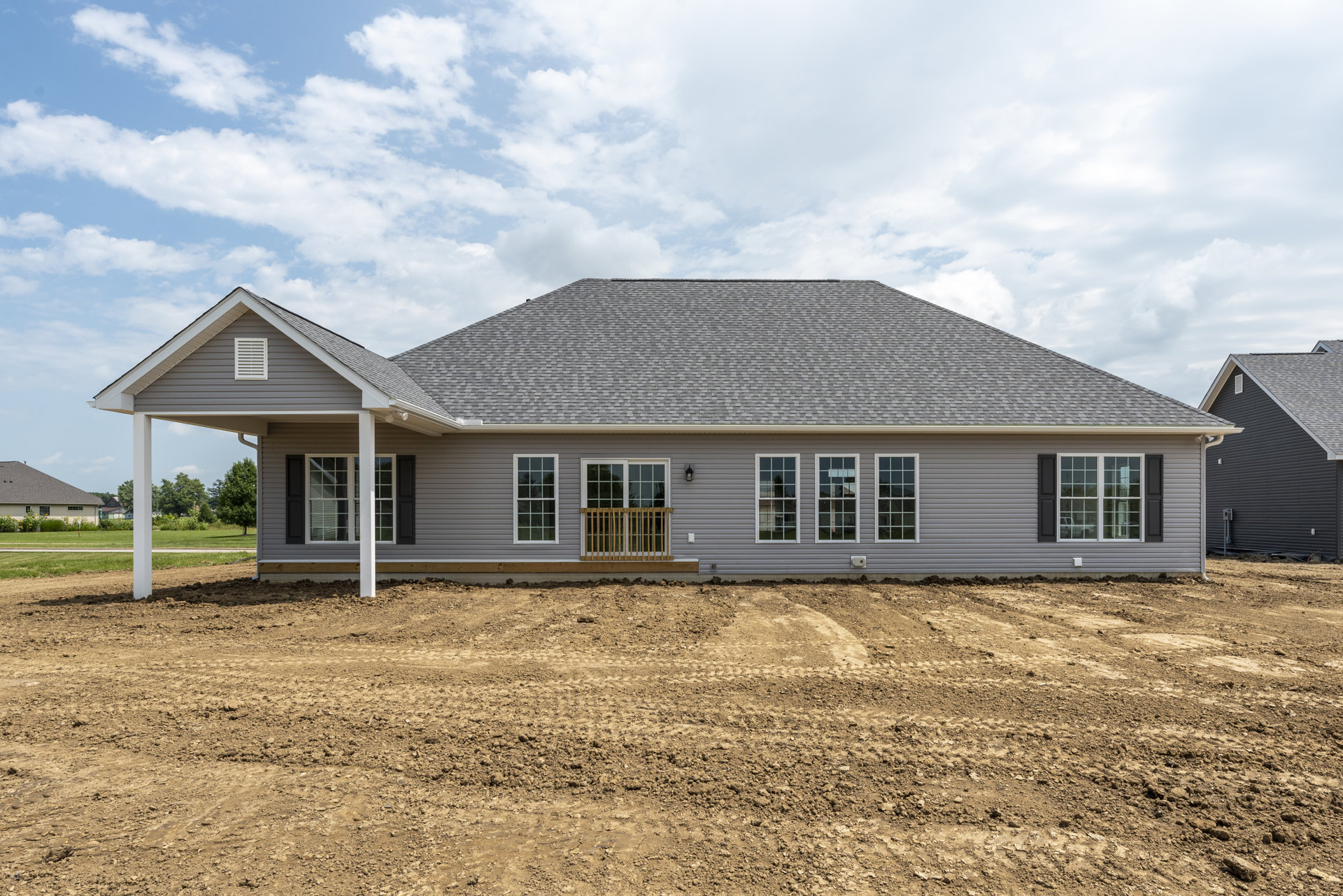 Black siding custom home with multiple square-paned windows, white frames, wooden railing, and expansive dirt field in front under a cloudy sky