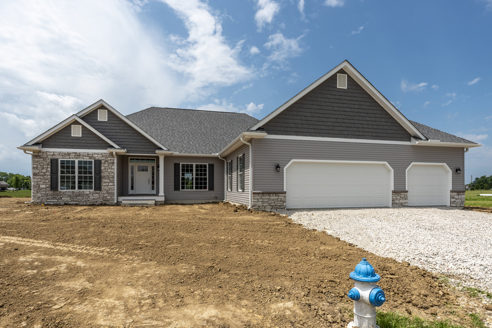 White garage door and entry door with glass panes, gray shingle roof, stone wall accents, blue and white fire hydrant on lawn, cloudy sky overhead