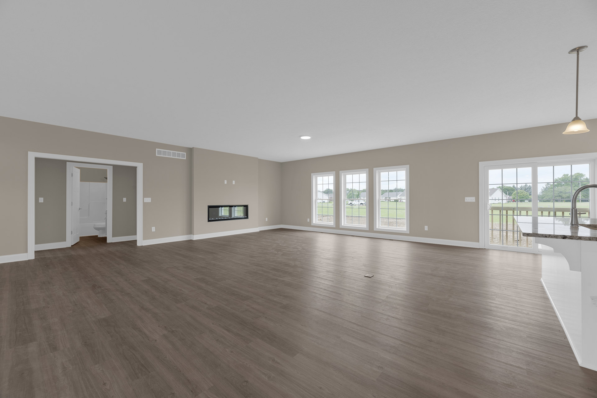 Spacious room with light wood flooring, tall windows overlooking a green lawn, white ceiling with recessed lighting, black-framed mirror, and doorway leading to adjacent space
