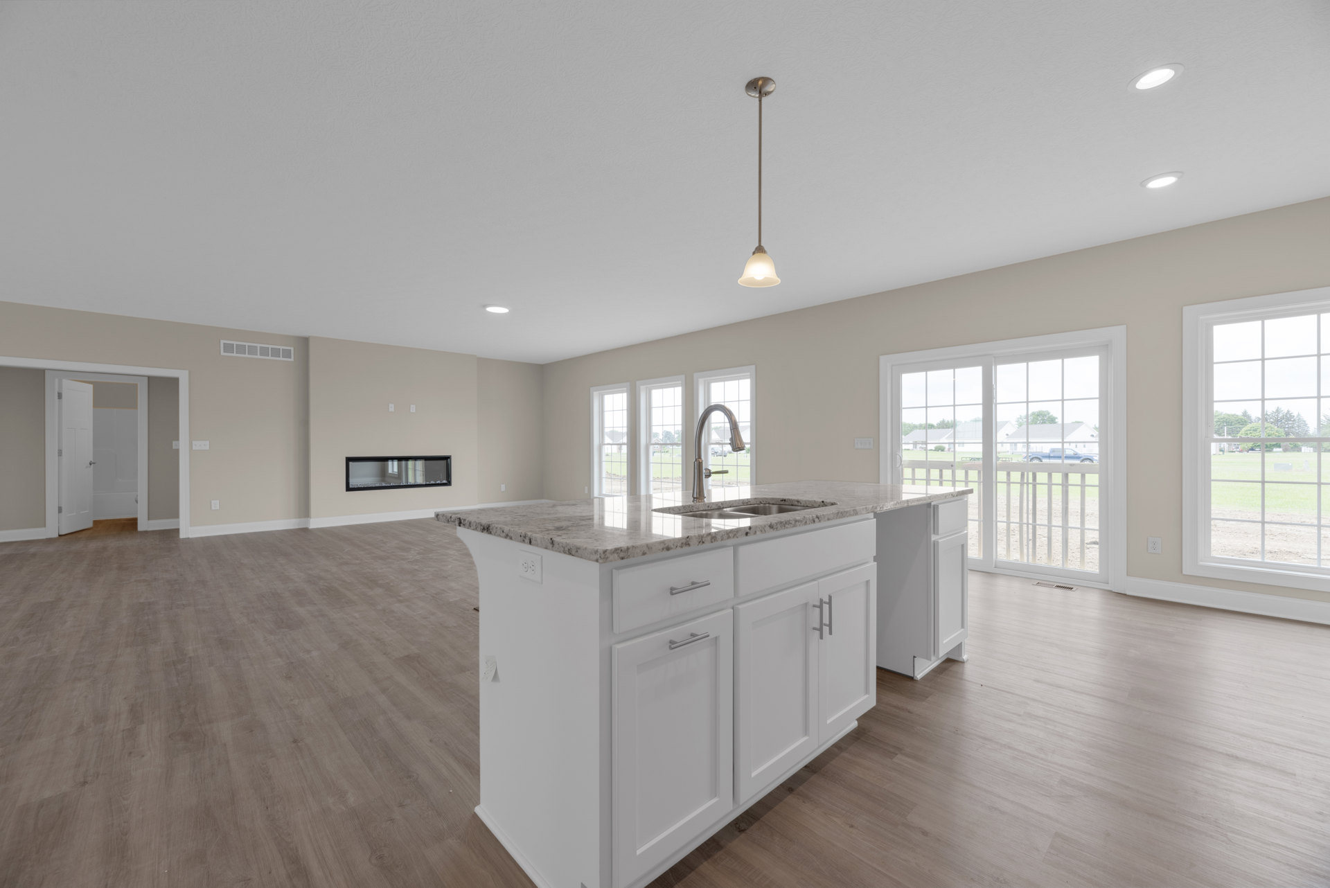 Spacious kitchen featuring a marble-topped island, sleek cabinetry, metal pendant lighting, large windows overlooking a grassy field and tree, and a wall-mounted mirror.