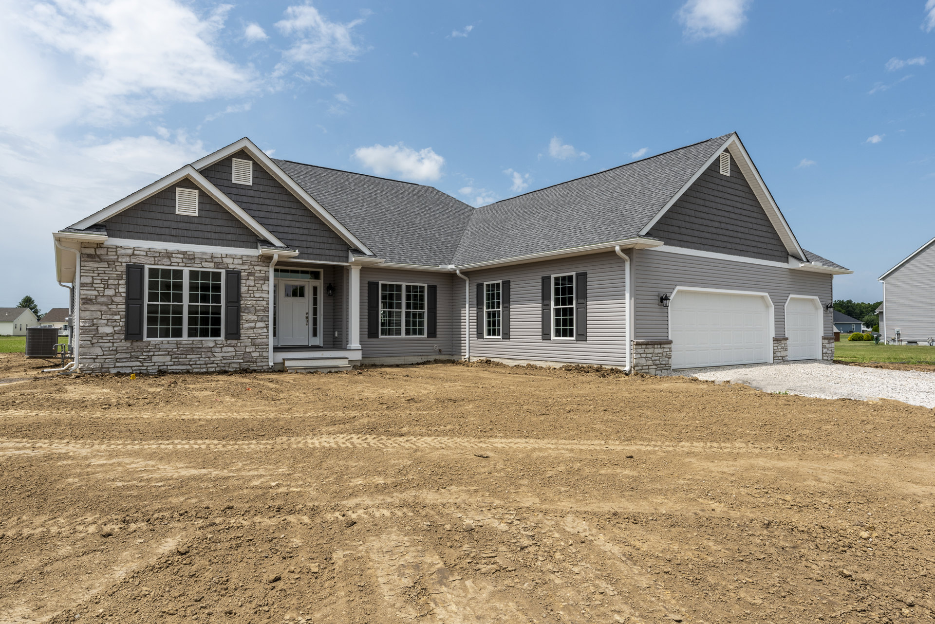 Two-story house with white garage door, grey roof, and white-trimmed windows, set beside a dirt field marked with tire tracks under a cloudy sky