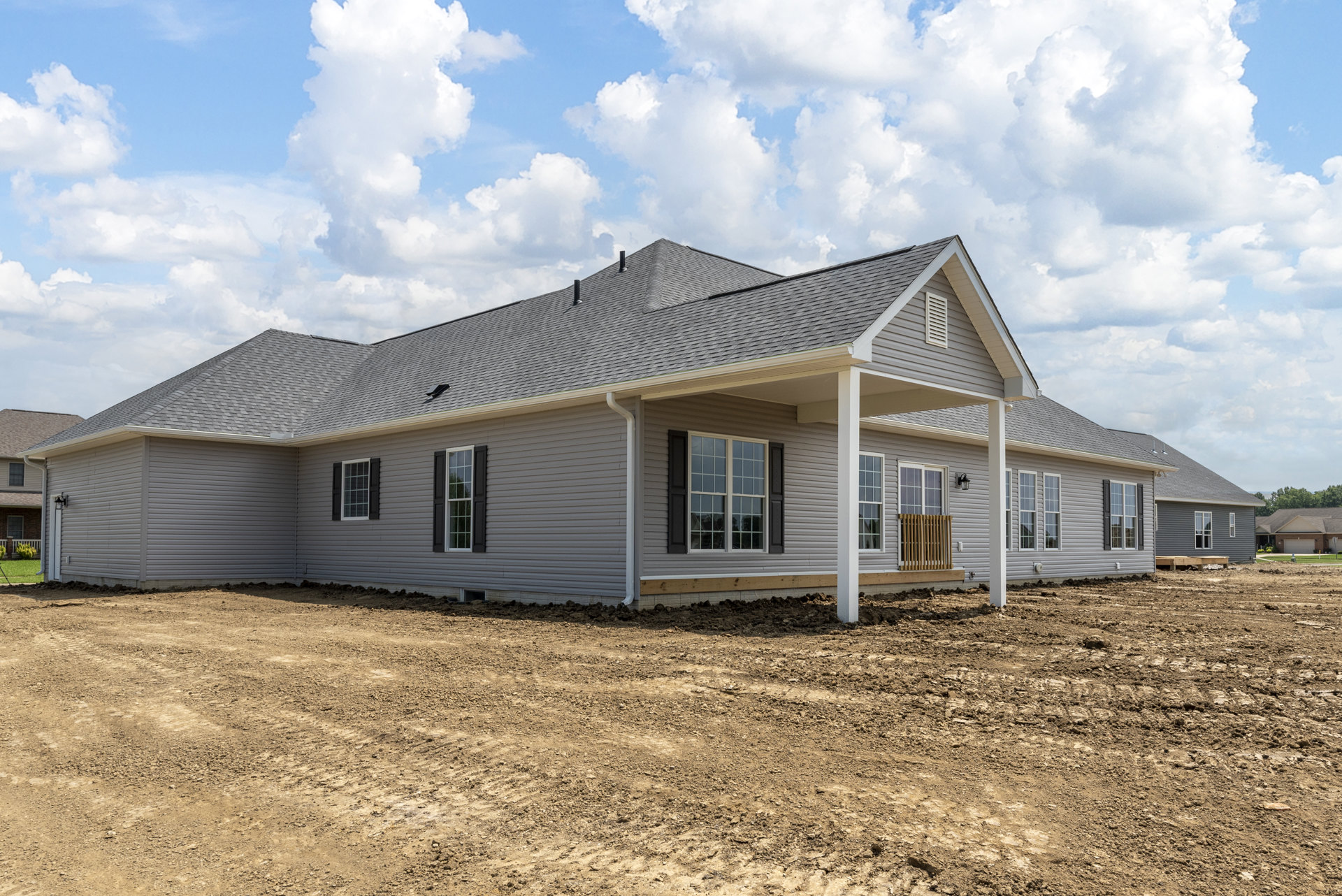 Partially built house with exposed wooden framing, black and white window shutters, dirt construction site, and cloudy blue sky overhead