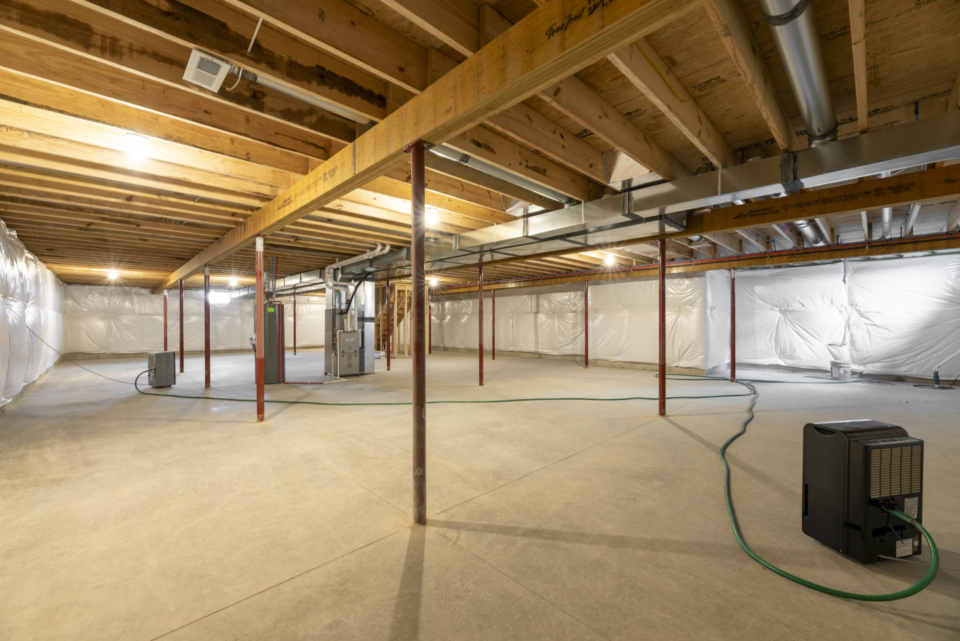 Exposed wooden ceiling beams above unfinished basement floor, white vent attached to wood surface, black machine with green hose and pipe, white fabric insulation visible on wall.