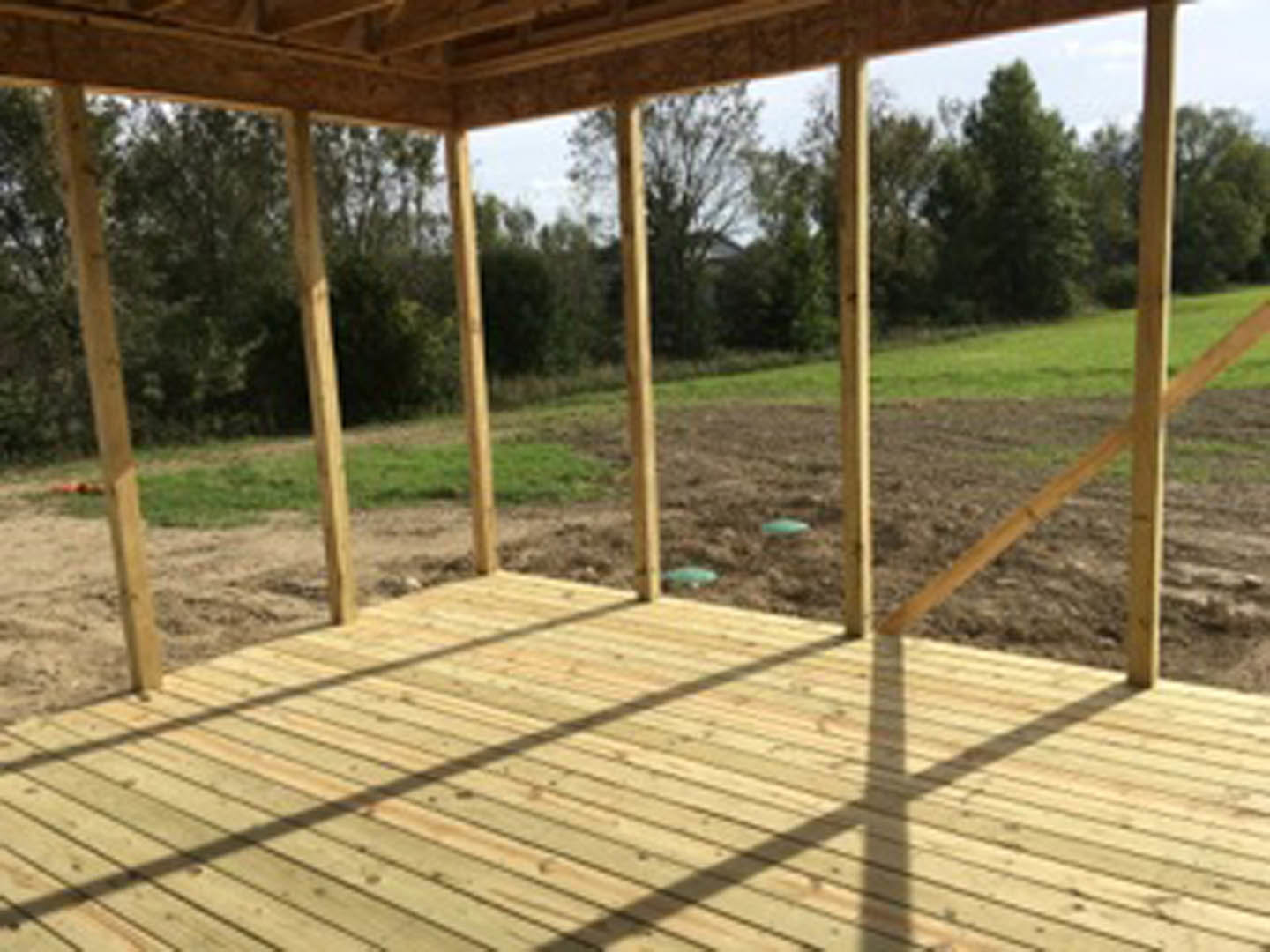 Wooden deck with plank flooring and railing overlooking grassy field bordered by mature trees under open sky