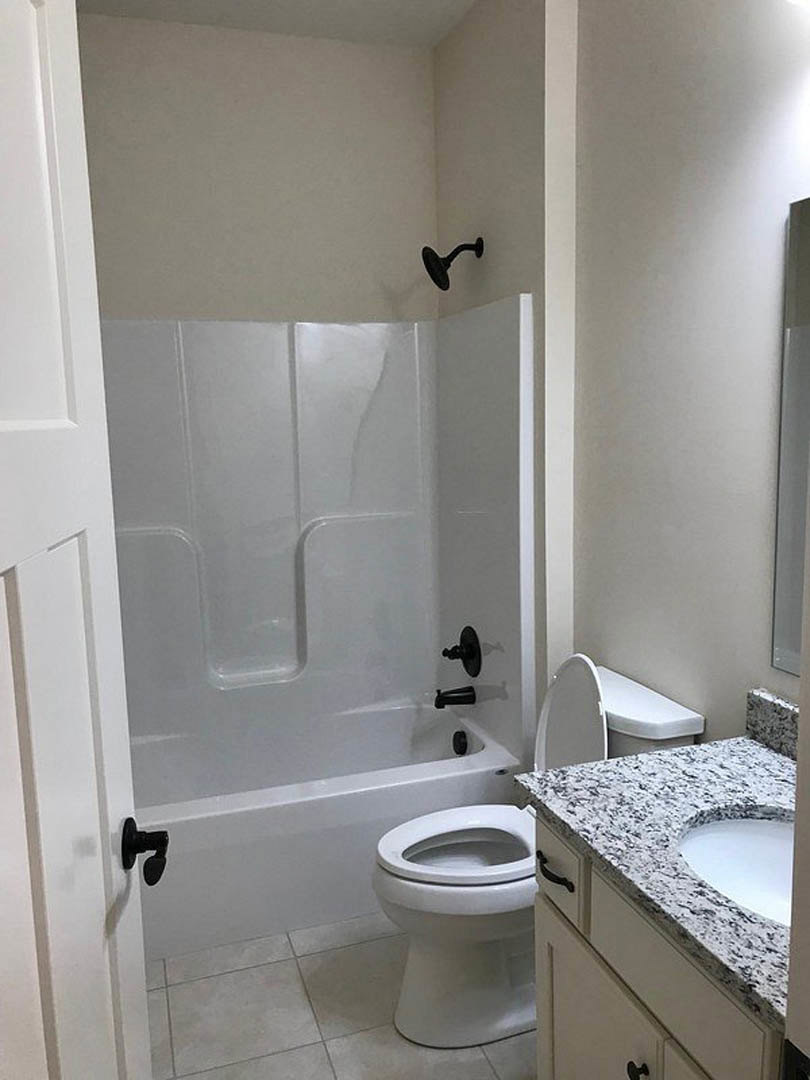 White porcelain sink set in a light countertop beside a modern toilet, with neutral tile flooring and partial view of a bathtub in a bathroom featuring simple fixtures and clean