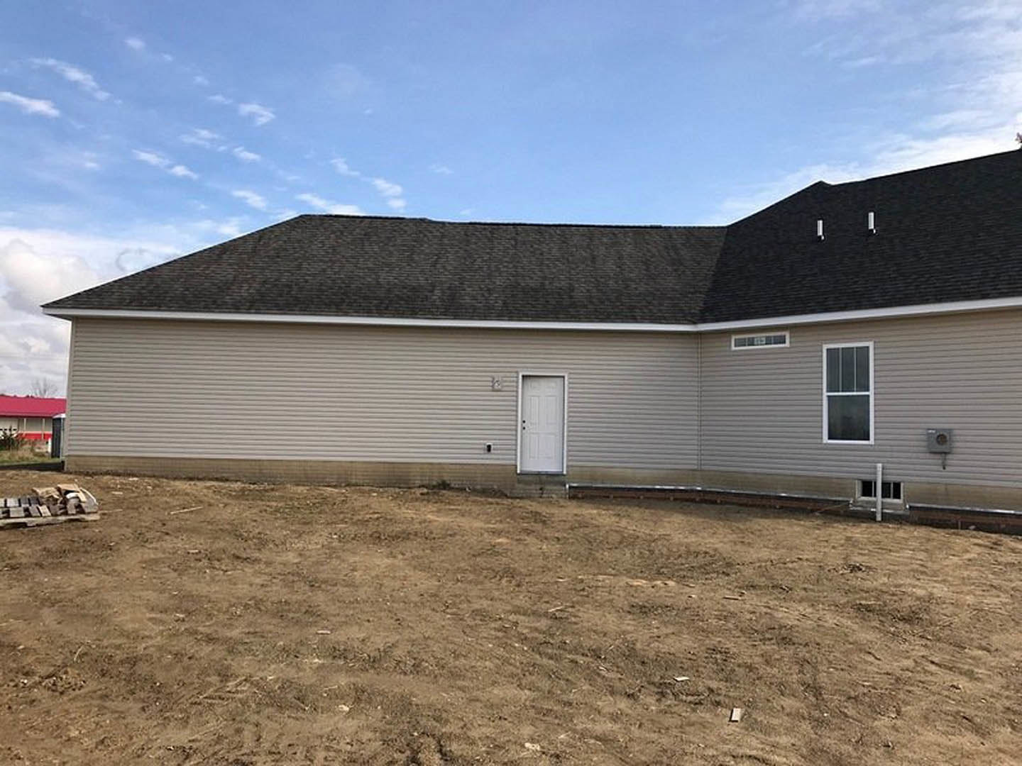 Red cottage-style home with white-framed windows and door, metal red roof, surrounded by a dirt field under a cloudy sky