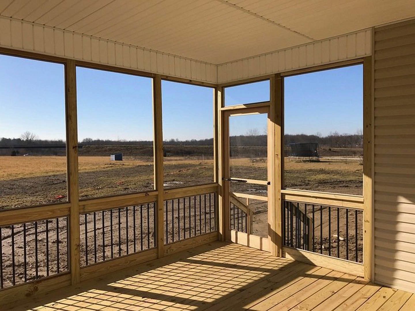 Wooden porch with metal railing overlooking open grassy field, large windows, and shaded deck area.