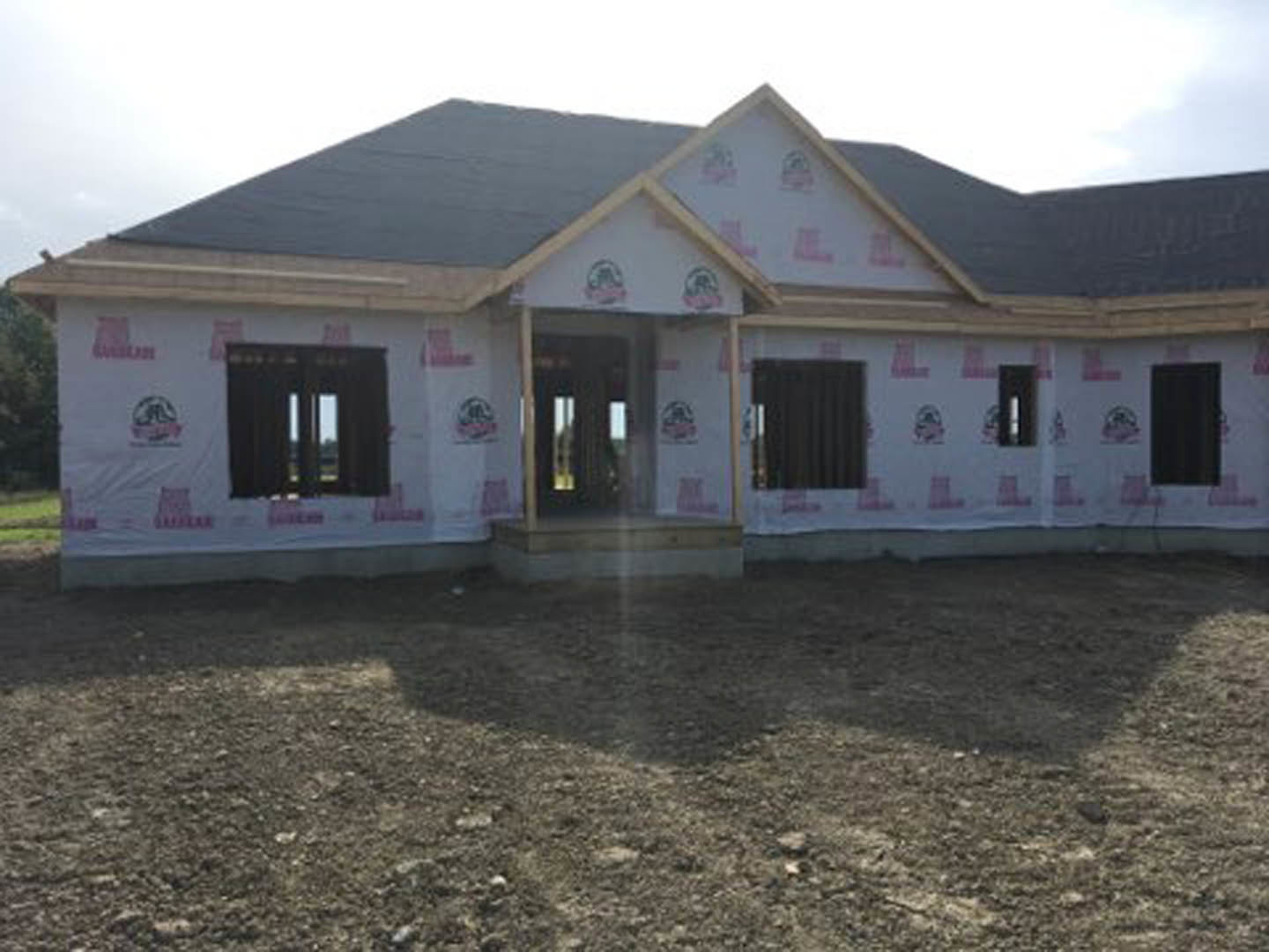 Partially built house with exposed framing, unfinished roof, red tape marking construction areas, dirt field in foreground, visible windows and door openings, cloudy sky overhead