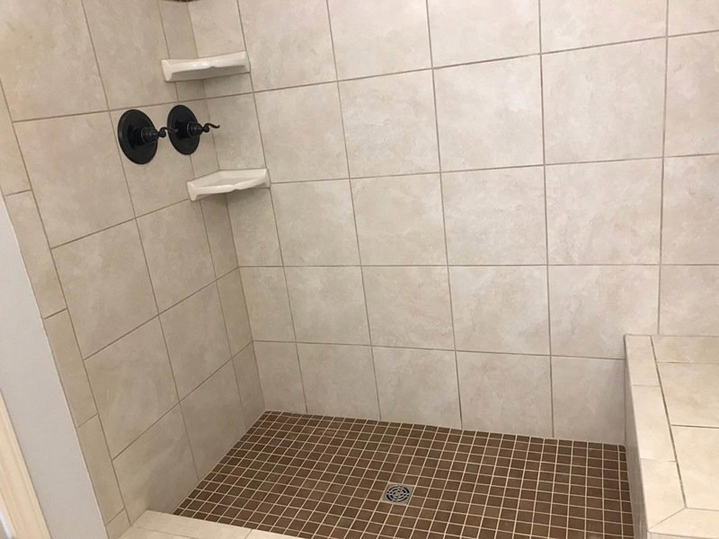 White tile shower with chrome shower head, linear drain, and adjacent white sink; black switches mounted on wall, clean white surfaces and edges throughout.