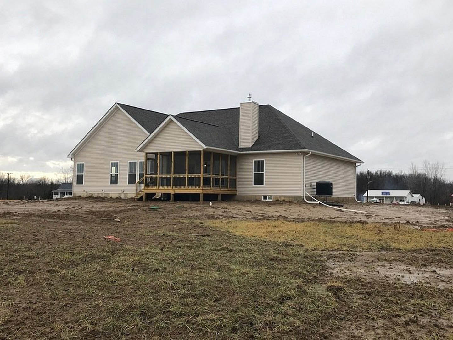 White farmhouse with black roof and covered porch, black wall-mounted box near entry, grassy lawn in foreground, Southfork Ranch visible in background under partly cloudy sky