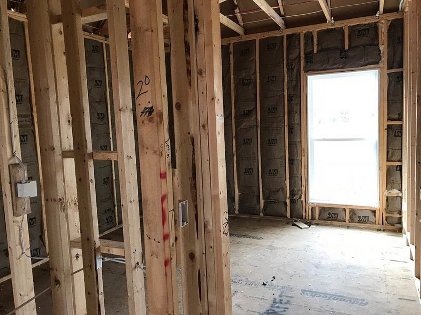 Wood-framed room under construction with exposed beams, concrete floor, window opening, and construction materials scattered throughout