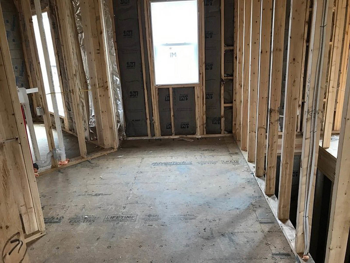 Unfinished room with exposed wood framing, concrete floor, window with white background, visible black pipe and construction materials