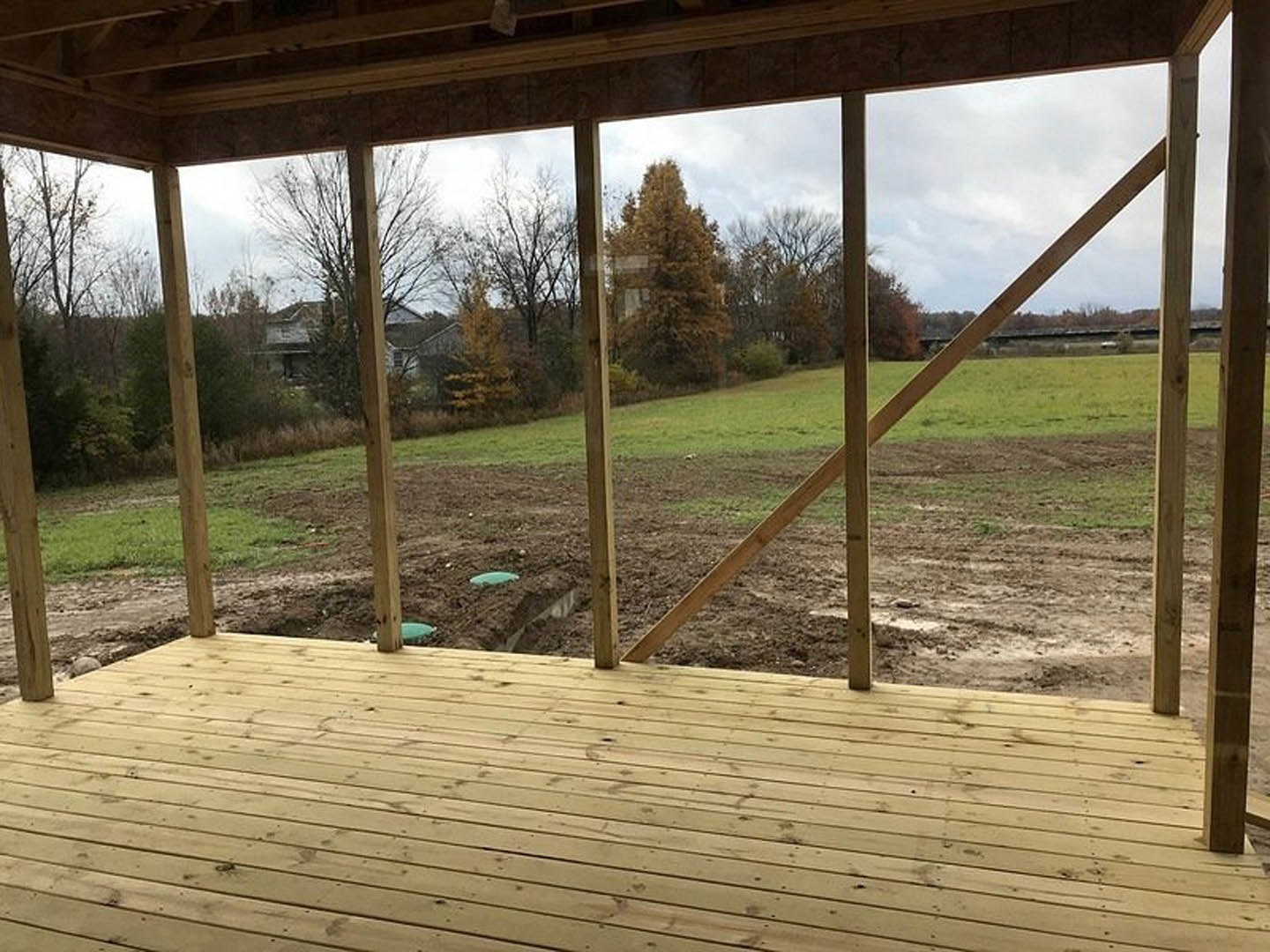 Wooden deck with railing overlooking a dirt field bordered by leafy trees, sunlight filtering through nearby window, patch of grass and green object visible in foreground.