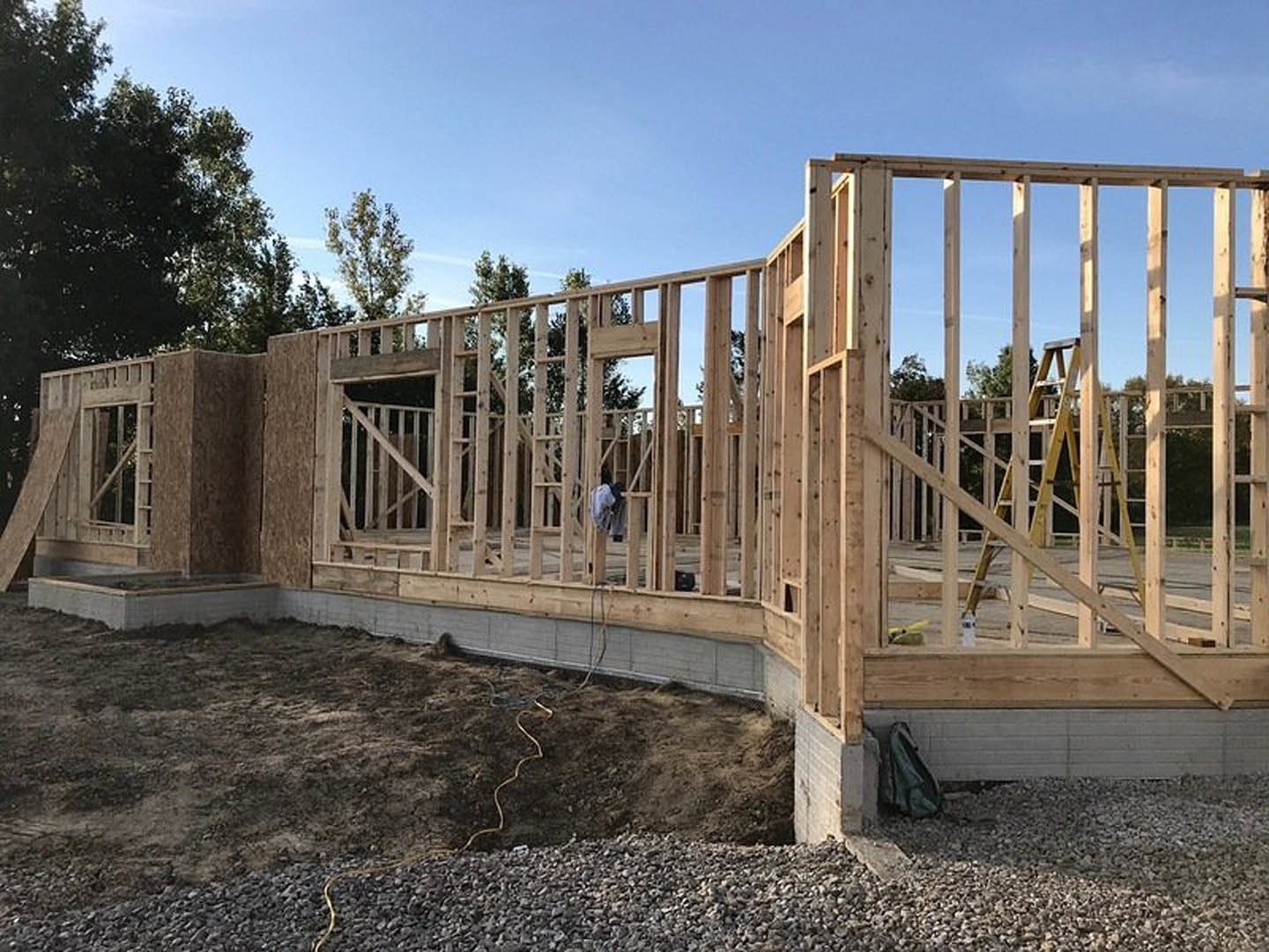 Wood-framed house under construction with exposed beams, ladder leaning against unfinished wall, person standing inside, dirt ground with yellow cord, blue sky overhead