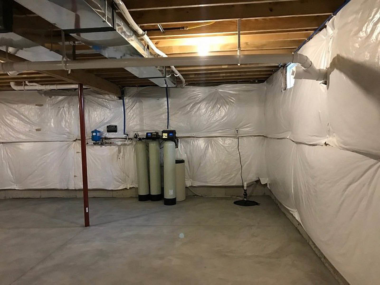 Unfinished basement with plaster walls, exposed ceiling, multiple pipes, cylindrical water tanks, white pillar with black border, and white object on concrete floor