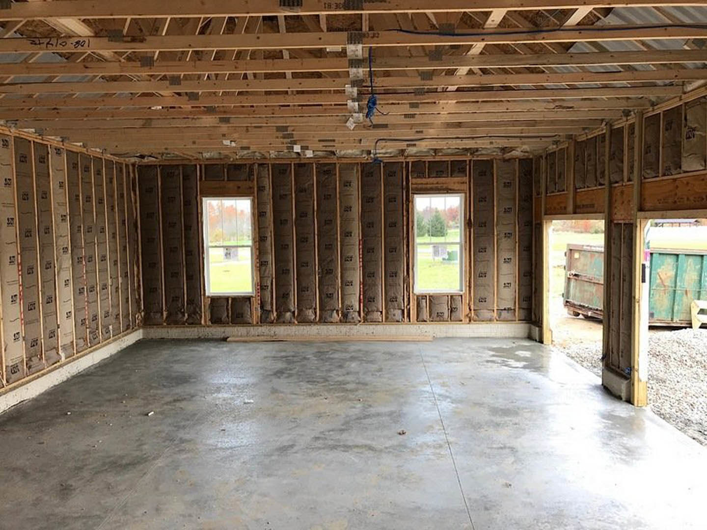 Concrete-floored room with exposed wooden beams, large windows overlooking trees and grass, blue electrical wiring, and a green electrical box on the floor