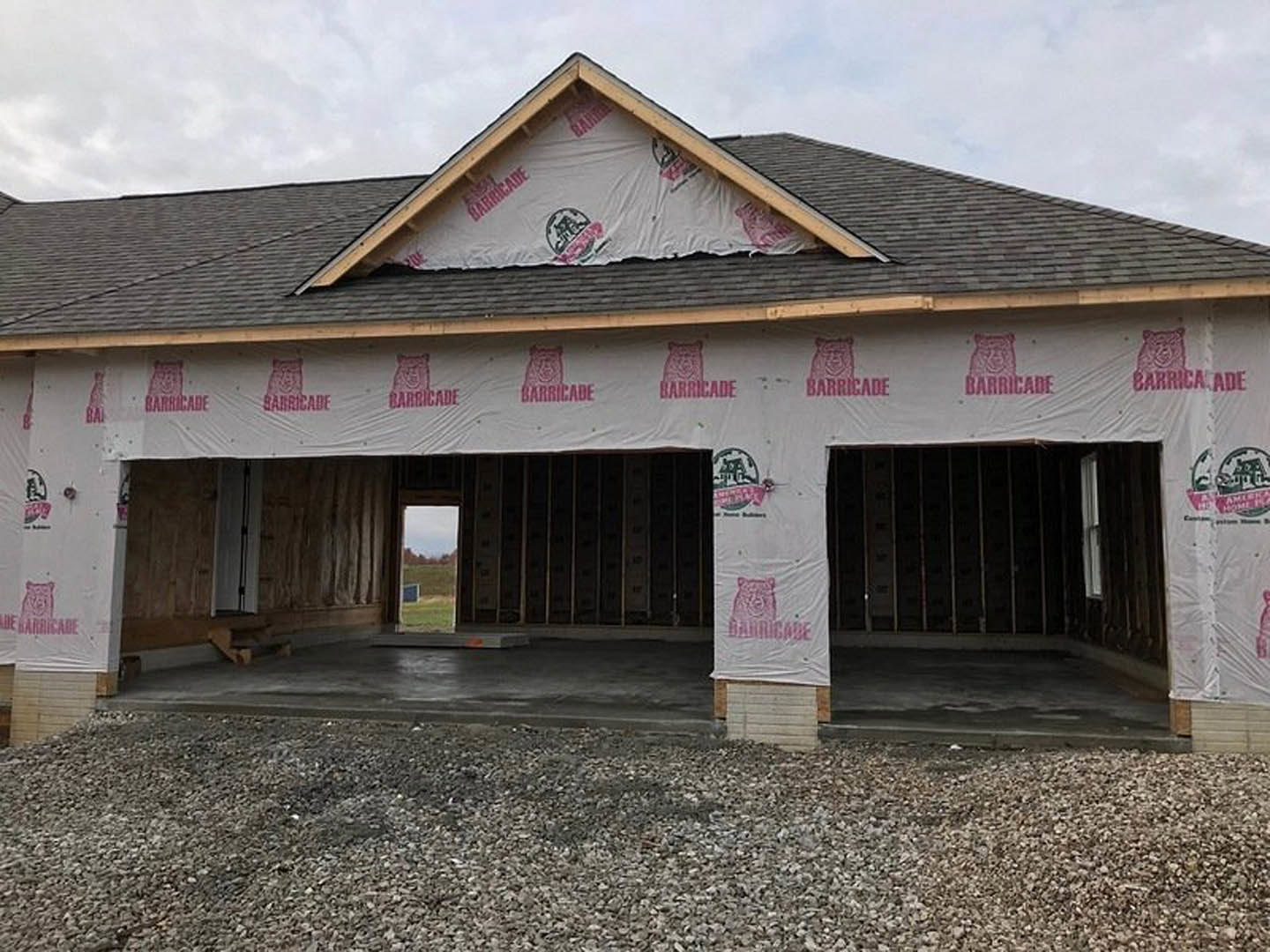 Partially built house with white tarp covering the roof, plastic sheeting over garage area, exposed gravel floor, metal siding, and window overlooking grassy field