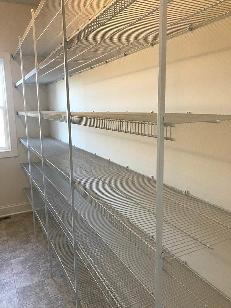 White plaster walls with empty steel shelves, tile flooring, window overlooking fenced field, metal rack mounted on ceiling