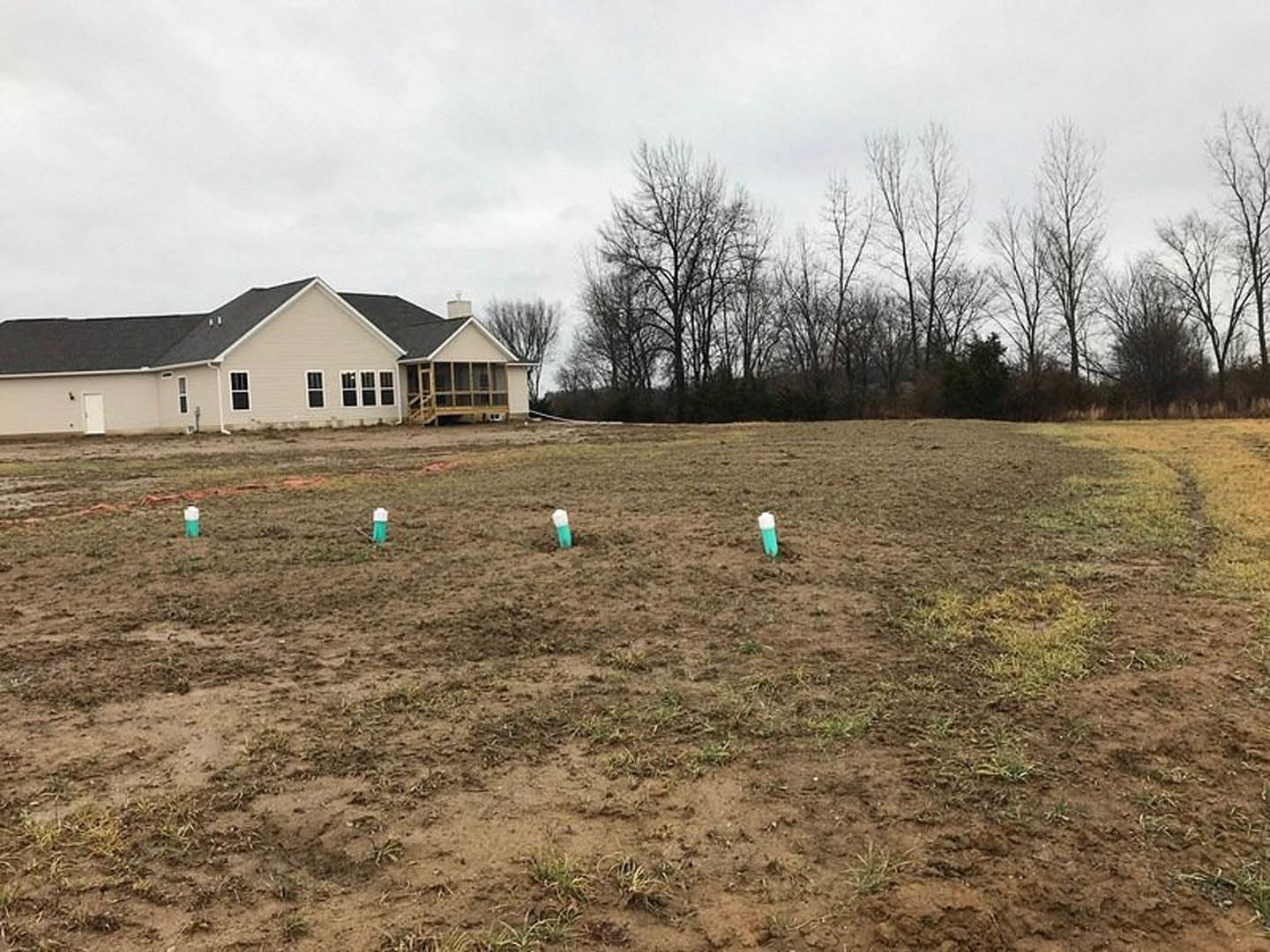 Modern house with black roof surrounded by a large dirt field, green poles scattered in foreground, leafless and leafy trees in background under partly cloudy sky