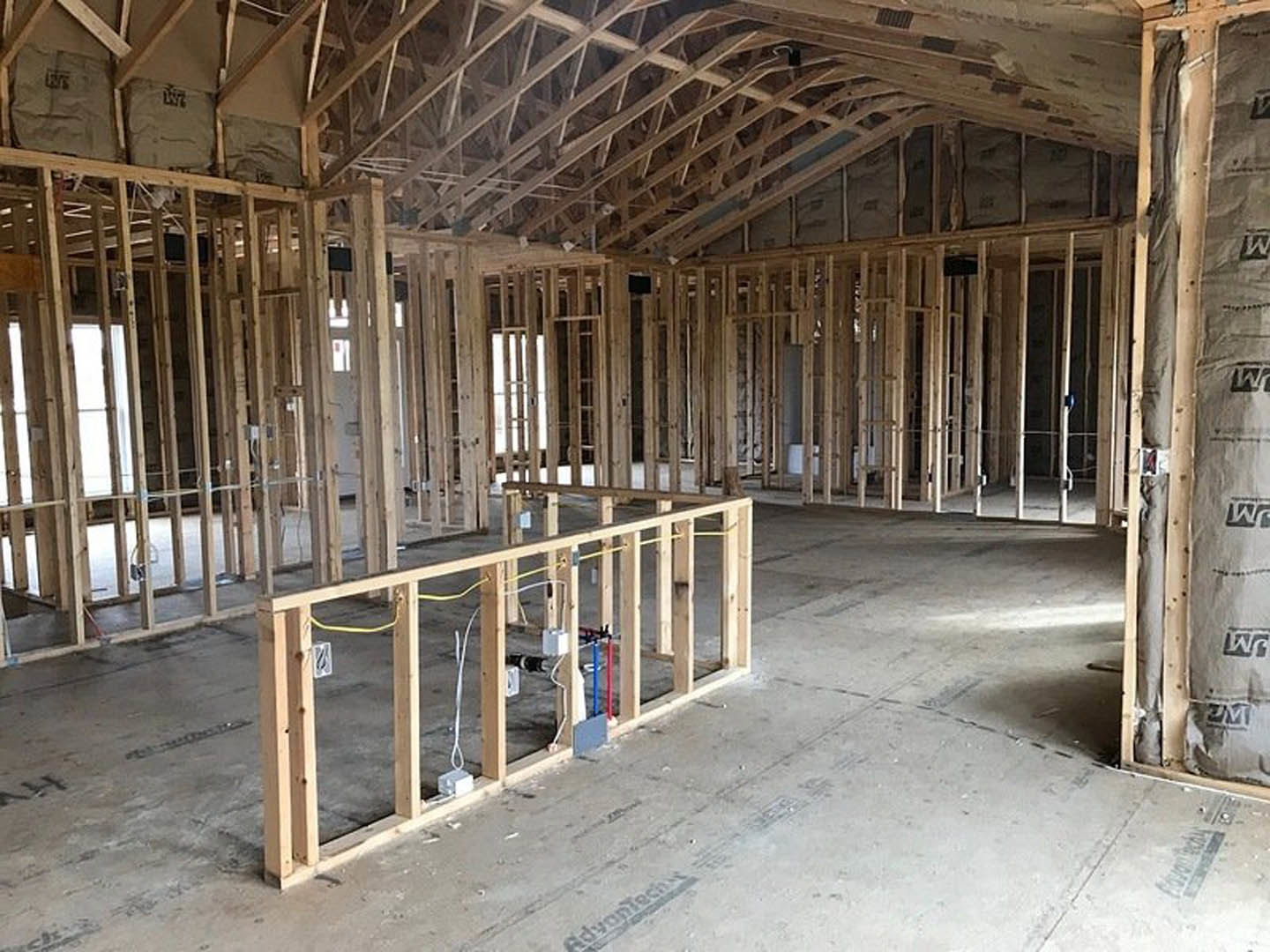 Exposed wood framing and roof trusses inside a house under construction, unfinished walls and ceiling with visible beams and floor structure