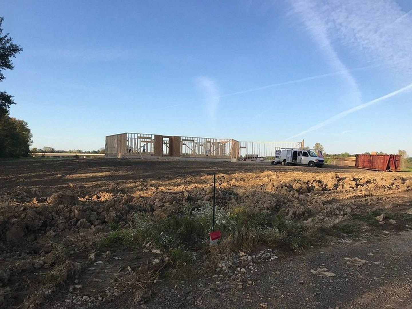 White work truck parked on dirt field near scattered grass, open van doors visible, tree with red marker in background, blue sky with clouds overhead, utility pole and train track